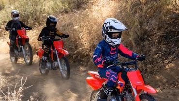Kids riding red dirt bikes on a dusty trail in protective gear.