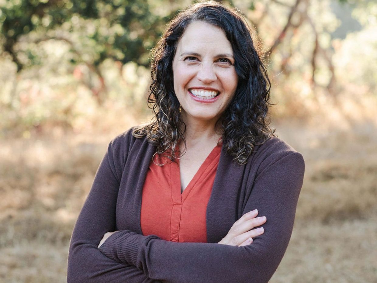 Smiling woman with curly hair wearing a brown cardigan and orange top outdoors.