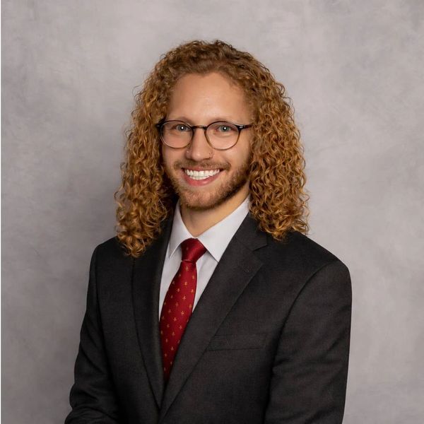 Smiling man with curly hair, glasses, and a suit with a red tie.