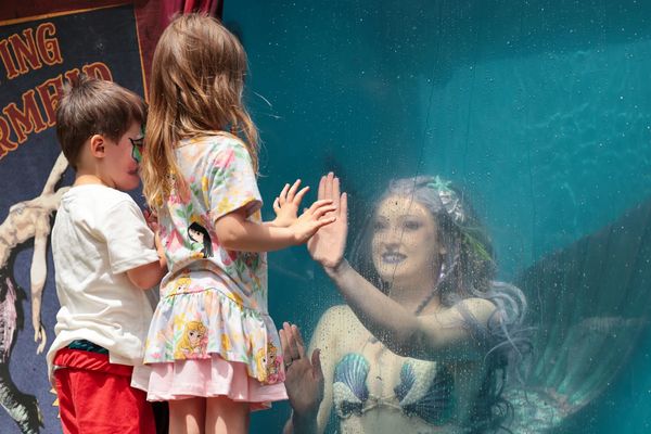Two children interact with a mermaid behind a glass panel at an event.