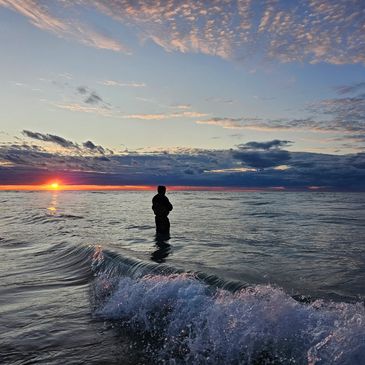 A person stands in the ocean watching a vibrant sunset with waves crashing nearby.
