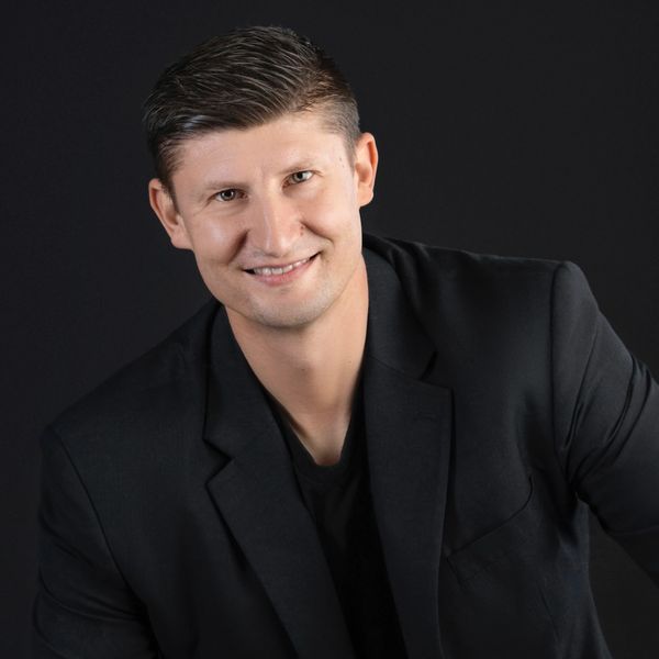 Smiling man in black blazer against dark background.