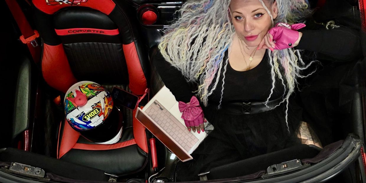 Woman with colorful braided hair inside a red Corvette, wearing pink gloves.