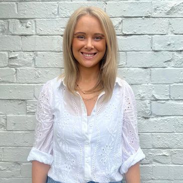 Woman smiling in white blouse and blue jeans against a white brick wall.