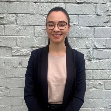 Young woman with glasses smiling against a gray brick wall.