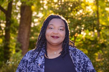 Smiling woman with braided hair in a floral shirt outdoors with sunlit foliage background.