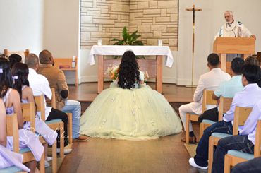 A girl in a beautiful gown kneels at an altar during a religious ceremony.