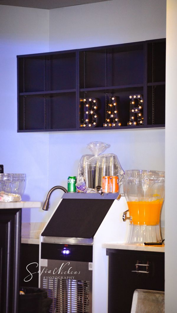 Illuminated BAR sign on black shelves above a drink station with soda and juice dispenser.