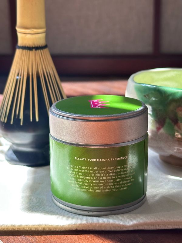 A green tin of Empress Matcha with a whisk and ceramic bowl on a cloth.