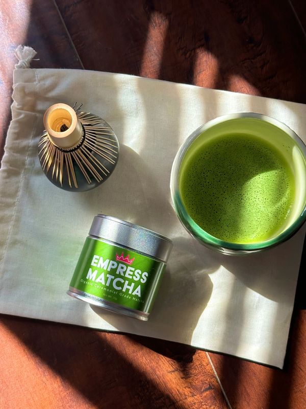 A cup of frothy matcha with a whisk and Empress Matcha tin on a cloth.