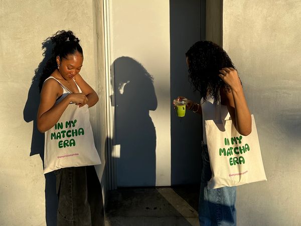 Two women with curly hair holding matcha-themed tote bags and a drink, standing in warm sunlight.
