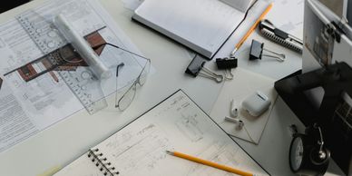 Workspace with architectural plans, sketches, stationery, and gadgets on a white table.