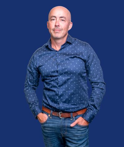 Confident bald man in a patterned navy shirt and jeans against a blue background.