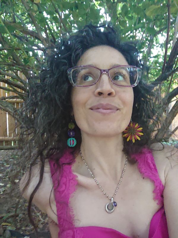 picture of donia under a tree. they have on a pink sleeveless top, glasses, curly hair, earrings.