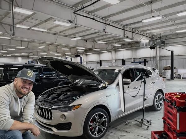 Man in a cap smiles while working on a white car with its hood open inside a garage.