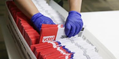 Image of mail-in ballots in a box being sorted by a poll worker.