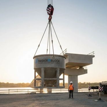 Construction worker overseeing a heavy concrete bucket lifted by a crane at a bridge site.