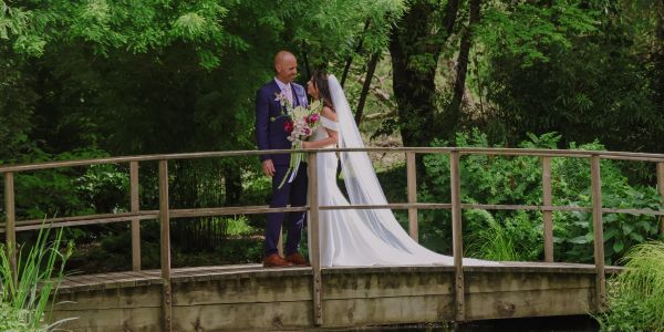 Newlywed couple standing on a wooden bridge surrounded by greenery.