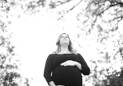 A pregnant woman in black holding her belly, looking upwards outdoors.