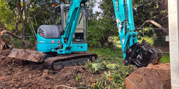 A teal Kobelco excavator parked on muddy ground with green foliage around.