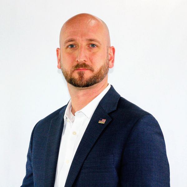 Man in a navy suit and white shirt with an American flag pin.