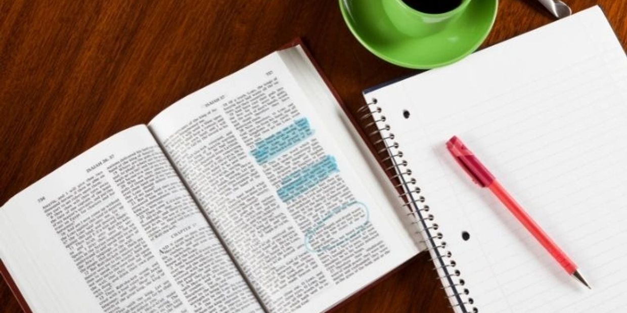 Open book, notebook, pen, and green coffee cup on wooden table.