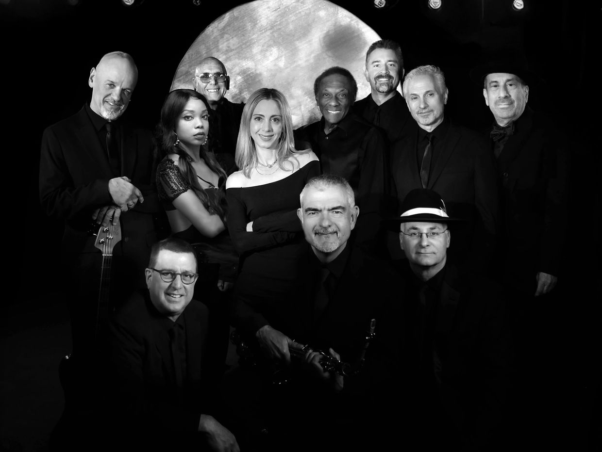 Black-and-white photo of a large, diverse band, The Dance Mechanics, posing with instruments in front of a circular backdrop.