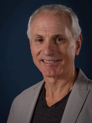 Smiling elderly man in a light gray blazer and dark shirt against a dark background.