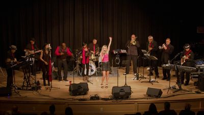 A lively band performs on stage with a female lead singer in pink.