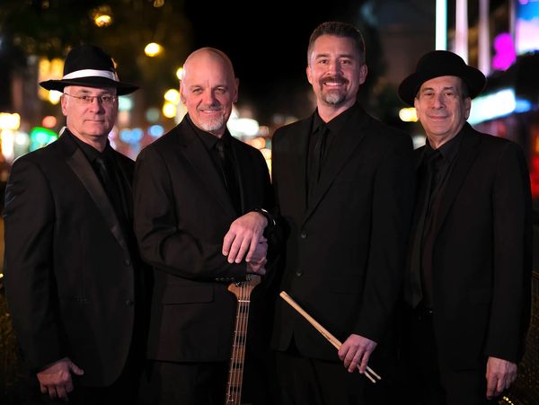 Four men who are members of the band The Dance Mechanics, are in black suits posing at night, two wearing hats, one holding drumsticks and another a bass guitar.There is a city backdrop behind them.