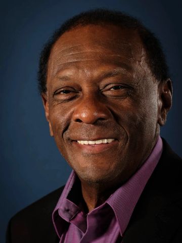 Smiling elderly man in a purple shirt and black jacket against a dark blue background.