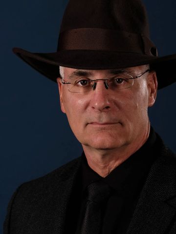 Mature man wearing glasses and a dark hat against a dark blue background.