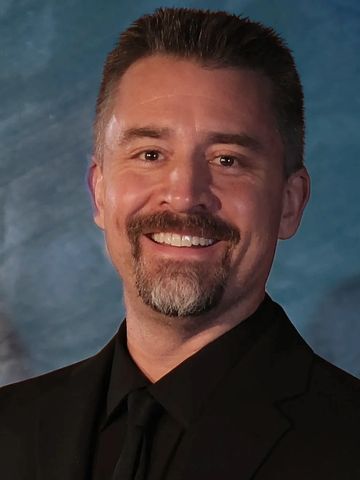 Smiling man with a trimmed beard in a black suit and tie.