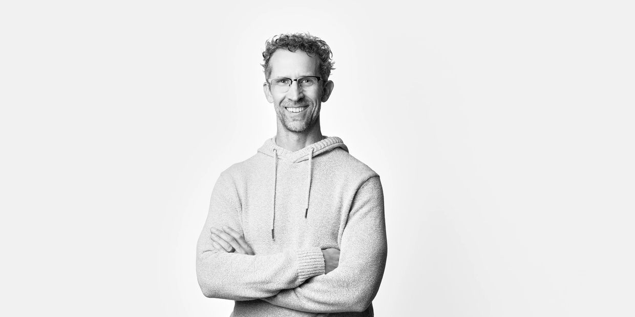 Smiling man with glasses in a light hoodie, arms crossed.