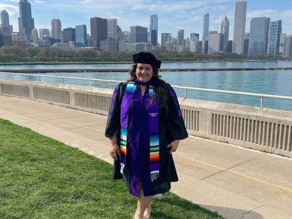 Graduate posing by a waterfront with a city skyline in the background.