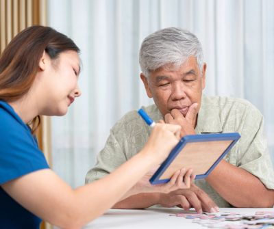 A caregiver explains something to an elderly man using a clipboard.