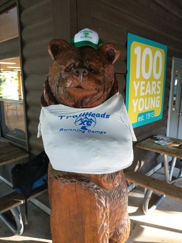 Wooden bear statue dressed in Trailheads Running Camps apparel near a 100 years young sign.