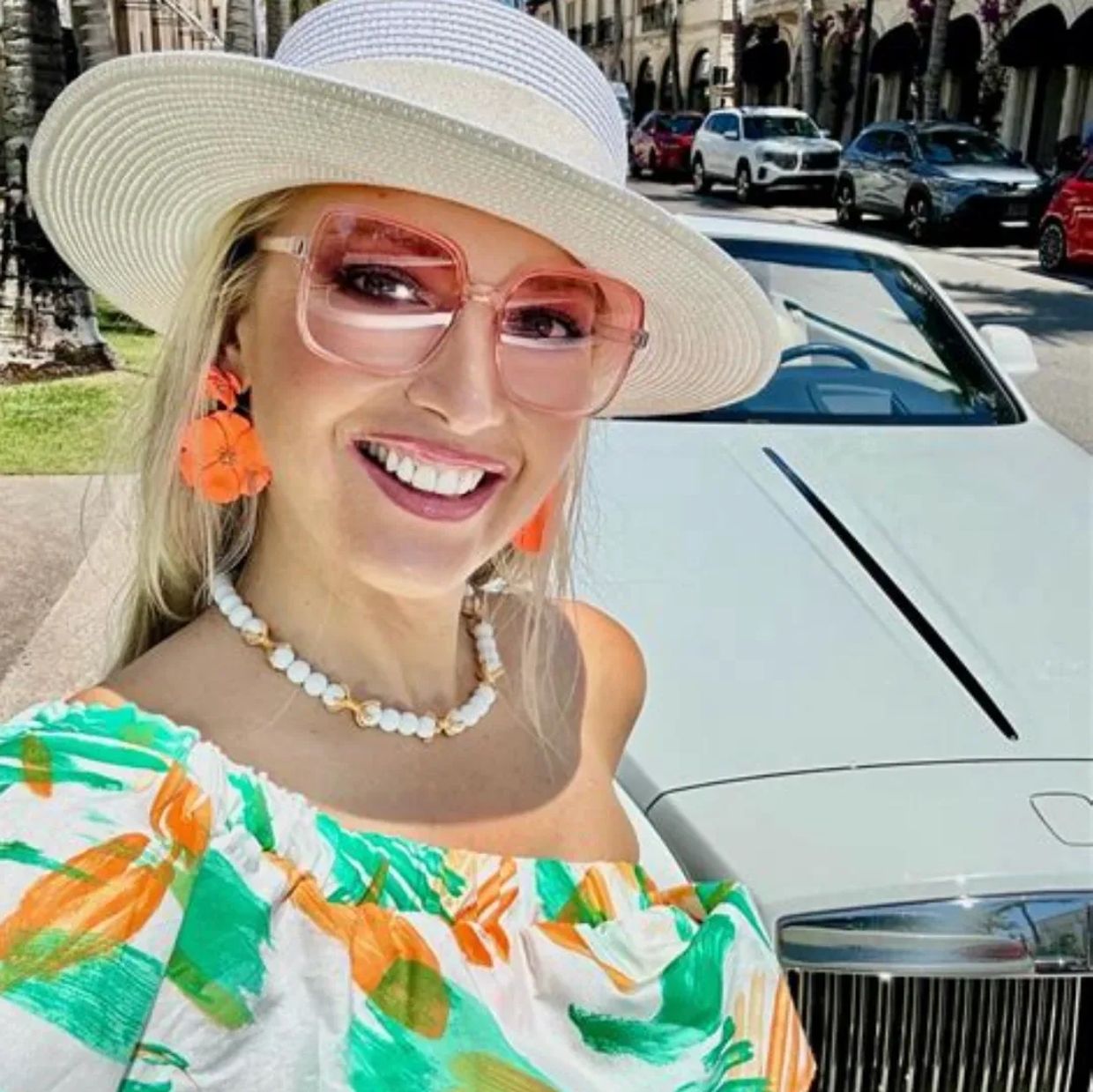 Smiling woman in a colorful dress and white hat poses in front of a luxury car on a sunny street.