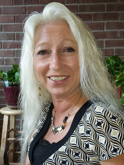Smiling woman with long white hair wearing a patterned dress and necklace indoors.