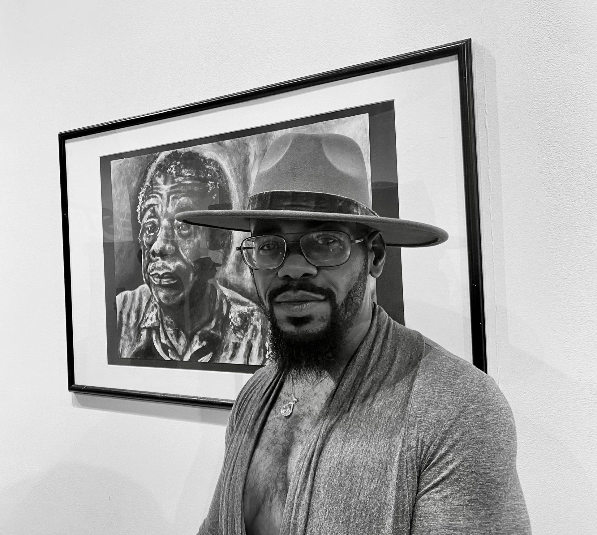 Man wearing a hat and glasses stands in front of a framed portrait painting.