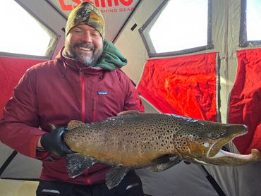 Smiling man holding a large brown trout inside a fishing tent.
