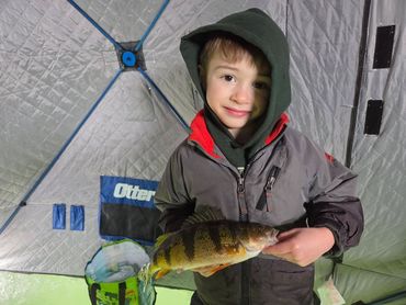 A young boy holding a freshly caught fish inside an ice fishing tent.