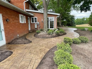 Curved stamped concrete walkway around a brick house with greenery and trees.