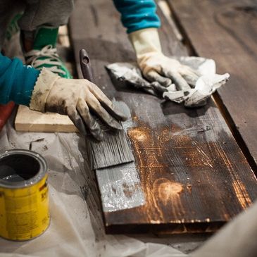 Person staining a wooden plank with a paintbrush while wearing gloves.
