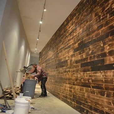 Worker using a saw to cut wood inside a room with a wooden accent wall.