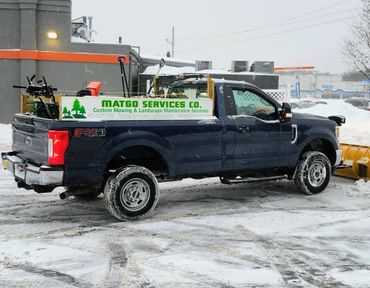 Blue pickup truck with landscaping equipment in snowy parking lot.