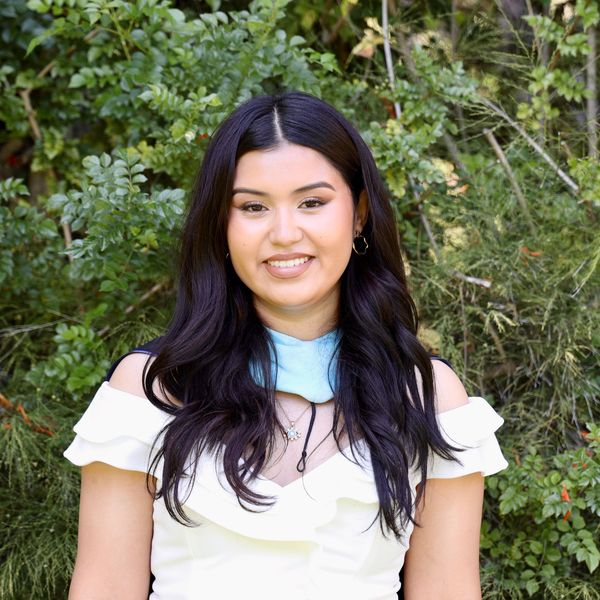Young woman smiling outdoors with long dark hair and white dress.
