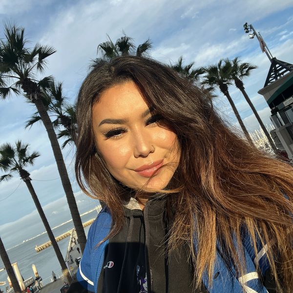 Young woman taking a selfie outdoors with palm trees and a waterfront in the background.