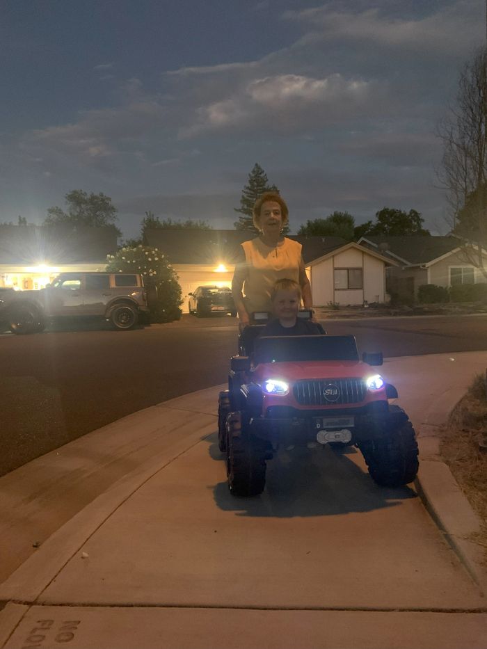 Child driving toy car with an adult pushing from behind at dusk.
