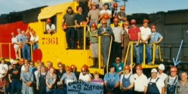 Group of railway workers posing around a yellow train engine with a congratulatory banner.